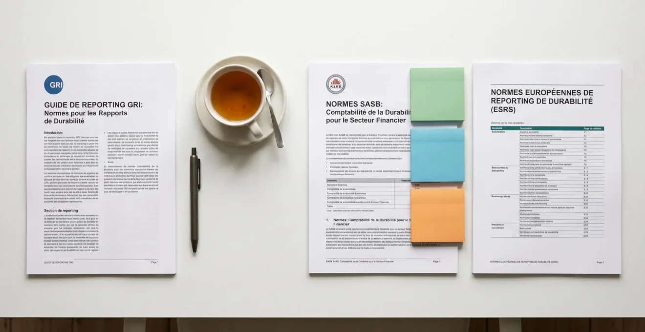 Vue de dessus montrant trois référentiels de reporting durabilité (GRI, SASB, ESRS) disposés côte à côte sur surface blanche avec stylo et post-it colorés, composition équilibrée et lumière douce